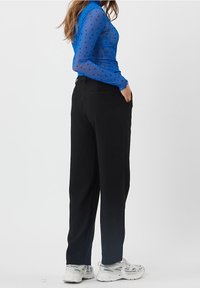 Sininen läpikuultava pitkähihainen paita, jossa on mustia sydänkuvioita, yhdistettynä korkeavyötäröisiin mustiin housuihin ja valkoisiin tennareihin, näkymä sivusta.