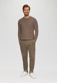Brauner strukturierter Strickpullover mit Rundhalsausschnitt, kombiniert mit hellbraunen Cordhosen und weißen Sneakers. Modell steht mit Händen in den Taschen.