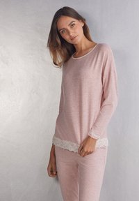 Lichtroze langwerpige top en bijpassende broek, gemaakt van een zachte stof. Opvallende kantafwerking langs de zoom en manchetten voor extra detail.
