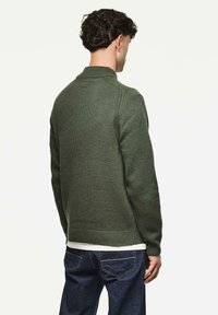 Mann mit dunklem lockigem Haar, der einen grünen Strickpullover und dunkle Jeans trägt, mit dem Rücken zu einem schlichten weißen Hintergrund gewandt.