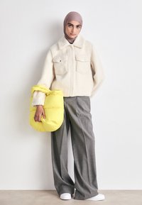 Cremefarbene Fleecejacke mit zwei Brusttaschen, kombiniert mit grauen gestreiften weiten Hosen. Hält eine leuchtend gelbe puffertasche und trägt weiße Turnschuhe.