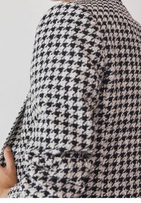 Gros plan sur le bras et le torse d'une personne portant une veste à motif pied-de-poule noir et blanc sur un fond uni.