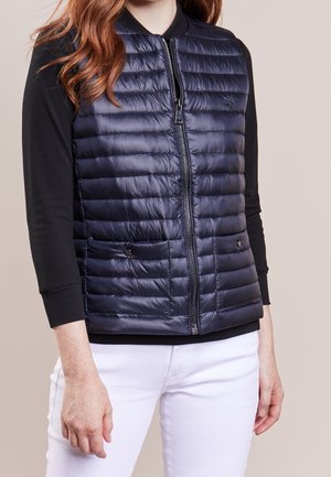 Frau trägt eine marineblaue, gesteppte ärmellose Weste über einem schwarzen Langarmshirt und weißen Hosen, steht vor einem schlichten Hintergrund.