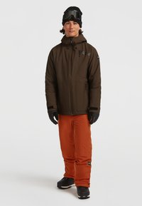 Sneeuwjack in donkerbruin met ritszakken, gecombineerd met oranje sneeuwbroeken en zwarte handschoenen. De outfit wordt aangevuld met een skibril en een gebreide muts.