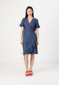 Robe en denim avec des manches évasées, décolleté en V profond et ourlet asymétrique. Présente un bord froncé et une couleur bleu foncé uni. Chaussures rouges et sac à main blanc.