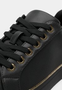 Gros plan sur une basket noire avec des lacets noirs, des œillets dorés et une bande dorée le long du côté de la semelle.