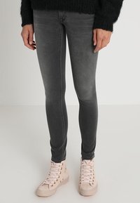 Persona con jeans ajustados negros desgastados y zapatillas altas rosa claro, de pie contra un fondo liso claro.