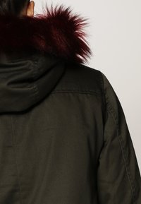 Osoba odwrócona plecami, ubrana w ciemnozieloną kurtkę z kapturem i burgundowym futerkiem na kapturze, na jednolitym tle.