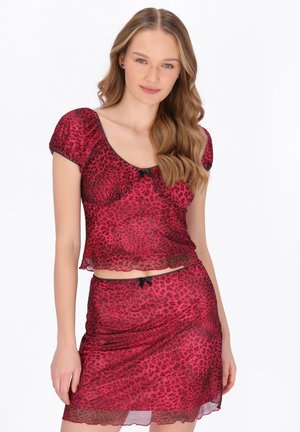 Młoda kobieta z długimi falującymi włosami, ubrana w przezroczystą bluzkę z krótkim rękawem w czerwono-czarne cętki i pasującą spódnicę, na białym tle.