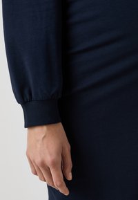 Námorně modrá dlouhá šaty s manžetou v ribu, vyrobené z hladkého materiálu. Detailní záběr ruky opřené o látku.