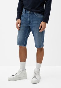 Jeansshorts mit einer ausgebleichten blauen Waschung, knielang geschnitten, fünf Taschen und Gürtelschlaufen, kombiniert mit weißen High-Top-Sneakern und hellen Socken.