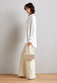 Vit skjorta med knappar, cremefärgade vida byxor, håller en guldglittrig kuvertväska med kedjerem. Naturligt trägolvsbakgrund.