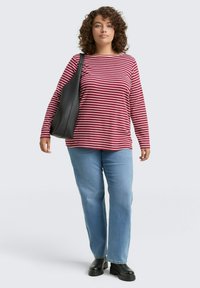 Vrouw met krullend haar, gekleed in een rood-wit gestreepte lange mouwen shirt, lichtblauwe jeans, zwarte laarzen en draagt een zwarte schoudertas.