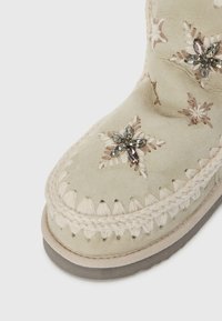 Beige ruskindsstøvle med blomsterbroderi, dekorative krystaller og flettet kant. Rund tå og fleksibel grå gummisål.