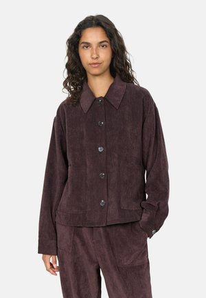 Femme aux longs cheveux bouclés portant une veste en velours côtelé violet foncé et un pantalon assorti, debout devant un fond clair uni.