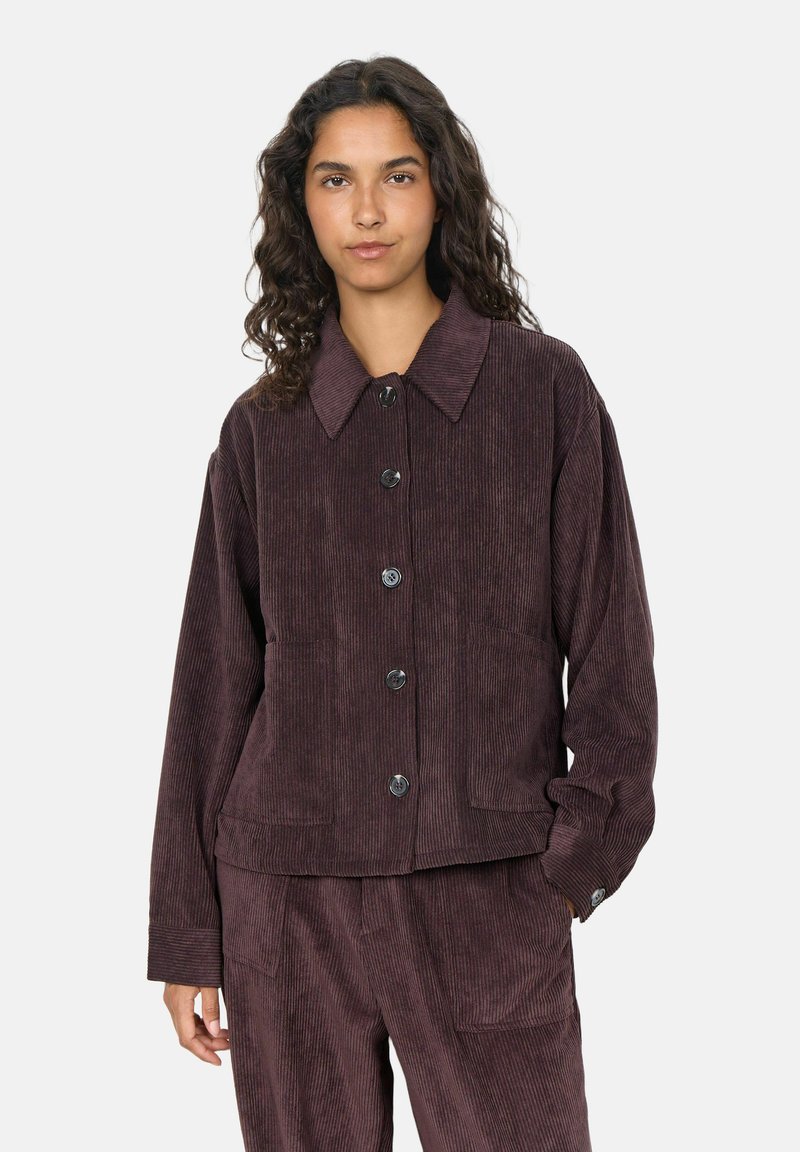 Femme aux longs cheveux bouclés portant une veste en velours côtelé violet foncé et un pantalon assorti, debout devant un fond clair uni.
