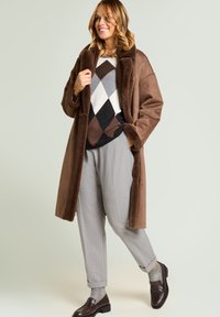 Abrigo de piel sintética marrón con un cuello ancho, combinado con un suéter estampado en gris, marrón y negro, y pantalones gris claro. Mocasines marrones.