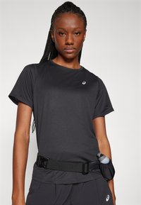 Donna con capelli intrecciati che indossa una maglietta sportiva nera e pantaloncini con una cintura in vita, che tiene in mano una bottiglietta d'acqua piccola.