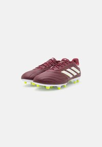 Scarpe da calcio color marrone scuro con tomaia sintetica testurizzata, tre strisce bianche e tacchetti giallo brillante. Design a profilo basso con chiusura a lacci.