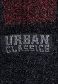 Juodas ir raudonas tekstūruotas audinys, kuriame išsiskiria ryškus pilkas „URBAN CLASSICS“ ženklas, išsiuvinėtas ant pleistro.
