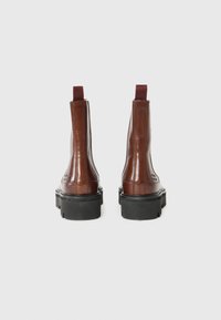 Bottines en cuir marron avec une finition brillante, panneaux latéraux élastiques et semelles en caoutchouc noir. Anses à l'arrière pour un enfilage facile.