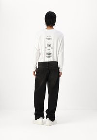 White long-sleeve shirt with graphic print on the back, paired with baggy black jeans and white sneakers, viewed from the back.