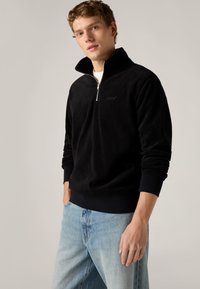 Μαύρο πουλόβερ από φλίς με φερμουάρ μέχρι τη μέση, ψηλό γιακά, ελαστικές μανσέτες και το λογότυπο της Levi's στο στήθος, συνδυασμένο με τζιν φως μπλε.