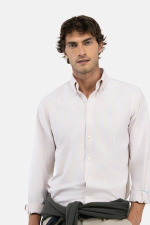 Hombre con cabello rizado y castaño lleva una camisa clara con rayas, abotonada, con las mangas remangadas y un suéter oscuro atado alrededor de su cintura.