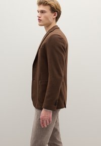 Braunes strukturiertes Sakko mit Kerbkragen, einer Tasche und Knopfdetails. Getragen über hellgrauen Hosen. Seitenansicht.