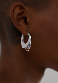 Sudraba apli auskari ar unikālu ovālu formu, kas satur trīs savstarpēji saistītus gredzenus un spīdīgu metālisku apdari, izceļot modernu dizainu.