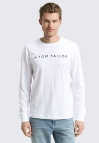 Junger Mann mit kurzen blonden Haaren, der ein weißes langärmliges Hemd von Tom Tailor und hellblaue Jeans trägt, steht vor einem einfarbigen Hintergrund.