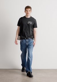 Μαύρο κοντομάνικο μπλουζάκι με λευκό κείμενο, μπλε τζιν παντελόνι και μαύρες δερμάτινες παπούτσια. Το μοντέλο κρατά μια ανοιχτό γκρι τσάντα, περπατώντας σε ουδέτερο φόντο.