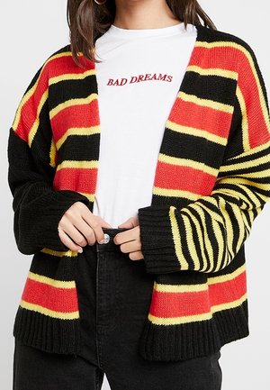 Femme portant un cardigan en maille rayé noir, rouge et jaune par-dessus une chemise blanche avec le texte rouge « BAD DREAMS », en train de fermer un pantalon noir.