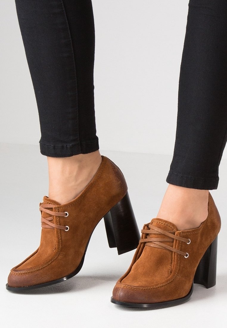 Zapatos de tacón alto de ante marrón con punta redonda, diseño con cordones y un tacón grueso negro. Acabado texturizado con detalles de costura en contraste.