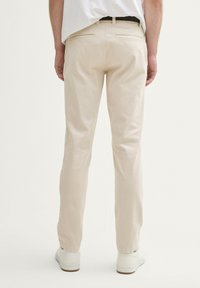 Personne vêtue de pantalons beiges, de baskets blanches et d'une chemise blanche, se tenant de dos contre un fond clair uni.