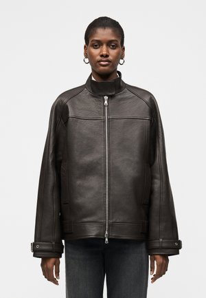 Γυναίκα που φορά μαύρο δερμάτινο μπουφάν με φερμουάρ, ψηλό γιακά, λουράκια με πόρπες στα φαρδιά μανίκια και σκούρο τζιν πάνω σε μονόχρωμο φόντο.