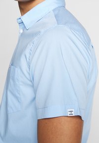 Ljusblå kortärmad skjorta med krage med knappar, vänster bröstficka och varumärkesetikett på ärmen. Slätt bomullstyg.