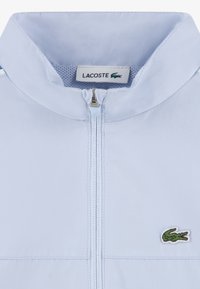 Ανοιχτό μπλε μπουφάν κατασκε υασμένο από λείο ύφασμα, με όρθιο γιακά, φερμουάρ μπροστά και πράσινο κεντητό λογότυπο κροκοδείλου.