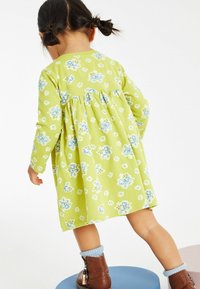 Ett litet barn med flätor, klädd i en grön blommig klänning, blå strumpor och bruna stövlar, som kliver ner från blå och beige runt plattformar.