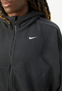 Czarna polarowa kurtka zapinana na zamek z kapturem, z wyraźnym białym logo Nike na piersi i teksturowanym materiałem. Widoczny zamek.