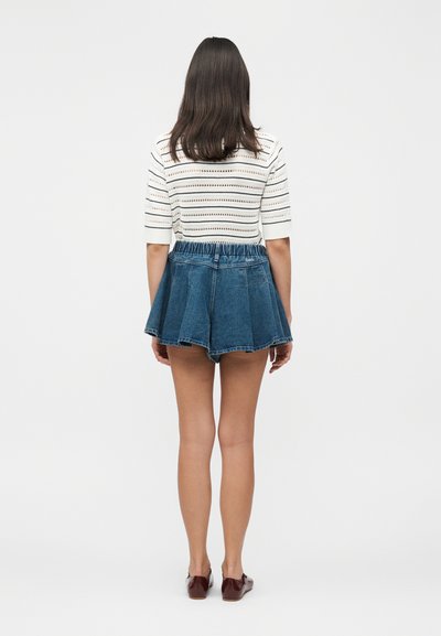 Γυναίκα που στέκεται με την πλάτη γυρισμένη, φοράει μια λευκή ριγέ κοντομάνικη μπλούζα, μπλε τζιν φούστα-σόρτς και καφέ επίπεδα παπούτσια σε μονόχρωμο φόντο.