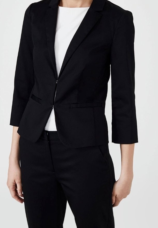 Femme portant un blazer noir cintré avec des manches trois-quarts par-dessus un haut blanc et un pantalon noir sur un fond uni.