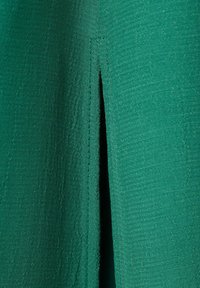 Detail tmavě zelené texturované látky s vertikálním průstřihem a viditelným šitím podél okrajů otvoru.