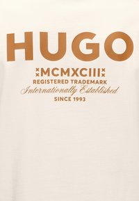 Λογότυπο HUGO με μεγάλα καφέ γράμματα πάνω από τα ρωμαϊκά ψηφία MCMXCIII, κείμενο «Καταχωρημένο εμπορικό σήμα», «Διεθνώς καθιερωμένο» και «Από το 1993» σε κρεμ φόντο.
