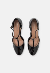 Scarpe Mary Jane in vernice nera con cinturino a T, punta tondeggiante e fibbie regolabili per la caviglia, viste dall'alto su uno sfondo bianco.