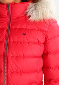 Czerwona kurtka puchowa o pikowanej fakturze, z futrzanym wykończeniem na kapturze i srebrnym zamkiem błyskawicznym. Małe logo na obszarze klatki piersiowej.