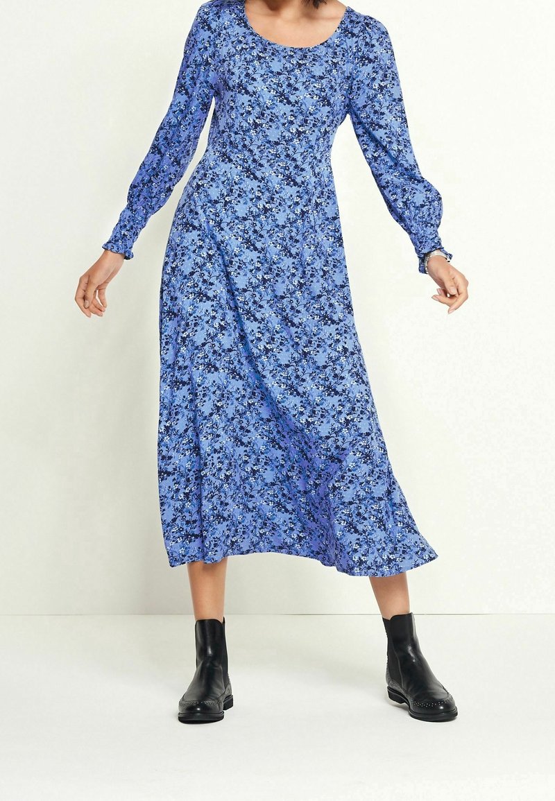 Femme portant une robe midi à manches longues à motif floral bleu, associée à des bottines noires, debout devant un fond clair uni.