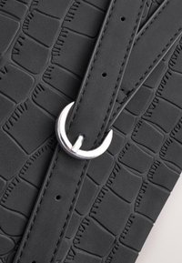 Melna teksturēta soma ar krokodila ādas rakstu, sudraba krāsas metāla sprādzi un gludu siksnu ar redzamām šuvju detaļām.