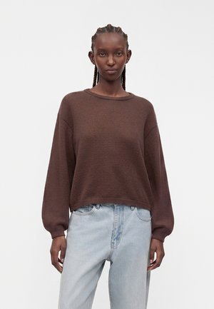 Kvinde med flettet hår iført en brun langærmet sweater og lyseblå højtaljede jeans, stående mod en ensfarvet lys baggrund.