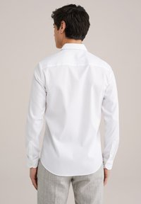 Bílá košile s dlouhým rukávem, s klasickým límečkem, manžetami na knoflíky a zaobleným lemem, vyrobená z hladké, lehké látky.